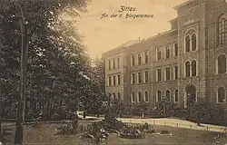 Die linke Hälfte der Hauptfront des heutigen Hauses 2 des Gymnasiums, drei Geschosse, fotografiert von schräg vor, davor mit Teilen der Parkanlage des Zittauer Stadtringes
