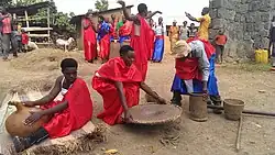 Auf einem unbefestigten Dorfplatz werden auf dem Boden verschiedene Gefäße vorbereitet. Rundherum stehen weitere, hauptsächlich junge, festlich rot gekleidete Menschen, die ihnen zusehen. Im Hintergrund sind einige Häuser und links ein kleiner Stall mit einem Rind und zwei Schafen.