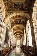 Loggia di Raffaello, die auf der rechten Seite offene Säulen und auf der linken Seite eine geschlossene Außenfassade mit Fenstern hat. An der Decke sind Malereien mit Rundbögen dazwischen, die die Säulen verbinden. Am Ende des Ganges steht eine Büste auf einem Podest.