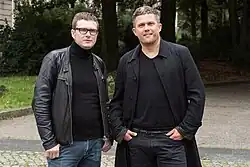Man sieht die beiden Moderatoren (links Ulf Buermeyer, rechts Philip Banse) auf einem gepflasterten Weg stehen. Im Hintergrund kann man ein bisschen Wiese und einen Wald erkennen