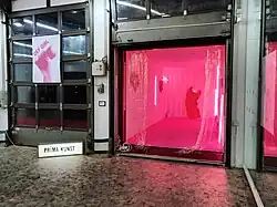 Das Bild zeigt einen Container, der an ein Liefertor gelagert wurde, sodass er das Foyer der Stadtgalerie Kiel erweitert. In dem Container befindet sich eine Ausstellung. Vor dem Container steht ein kleines Leuchtschild mit der Aufschrift Prima Kunst.