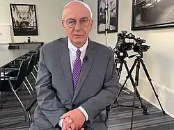 Ein Mann in grauen Anzug und rötlicher Krawatte mit einer dünnen Brille, Halbglatze und verschränkten Händen sitzt im Vordergrund. Im Hintergrund ist sind eine Stuhlreihe und zwei Videokameras auf Stativen zu sehen. An den Wänden hängen diverse Fotografien.