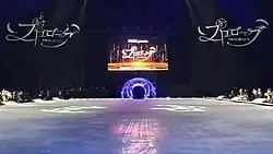 Blick über die angestrahlte Eisfläche auf den Künstlereingang und die darüber angebrachte Großleinwand, links uns rechts der Leinwand das Logo der Show, an den Seiten Ausschnitte des Publikums