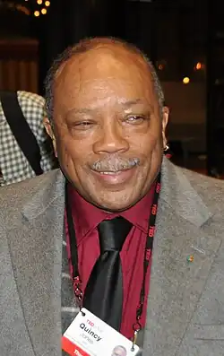 An older man with a white mustache, wearing a black tie, red dress shirt, gray jacket, and a name badge that hangs from a string around his neck.