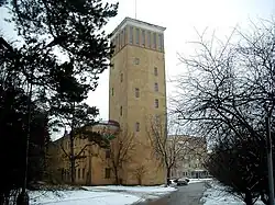 Gebäude von vorn mit Zuwegung, mit großem turmartigen mehrgeschossigen Bau in der Mitte, gerahmt von zwei flacheren ebenfalls mehrgeschossigen Bauten, sämtlichst in einem ähnlichen sandsteinfarbenen Ton in Farbe.