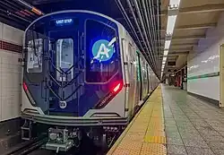 Zug am Bahnsteig eines U-Bahnhofs. Rechts der Bahnsteig mit weiß gefliesten Wänden, Bodenplatten aus grau gemustertem Betonwerkstein, Bahnsteigkante mit taktiler gelber Kontrastmarkierung. Links ein stehender Zug, silbergrau lackiert, dunkelblaue Frontelemente, alles noch neu und glänzend. Leuchtende LED-Anzeige mit blauem Liniensignet vorne, rot strahlende Rücklichter, oben Zielanzeige LAST STOP, an den geöffneten Seitentüren strahlen grüne LED-Streifen auf Bahnsteig und Wand.