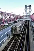 Blick vom Fußgängersteg auf die darunter verlaufende Brückenrampe der Williamsburg Bridge. Man blickt schräg von oben-vorne auf die Front und das Dach der ersten Wagen eines in gesicktem Edelstahl verschalten U-Bahn-Zugs. Die Brückenkonstruktion erscheint in einem bleichen Rotton, auf den Kraftfahrzeugfahrbahnen links und rechts ist wenig Verkehr sichtbar.