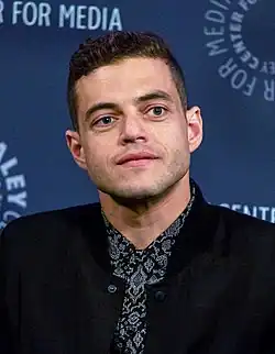 Rami Malek mit kurzen, dunklen Haaren und blauen Augen, der eine schwarze Jacke über einem gemusterten Hemd trägt, schaut nachdenklich. Der Hintergrund besteht aus einem dunkelblauen Banner mit weißem Text, der in einer kreisförmigen Anordnung den Namen „PALEY CENTER FOR MEDIA“ zeigt. Das Bild wurde bei einer Veranstaltung auf dem PaleyFest, New York 2015 aufgenommen.