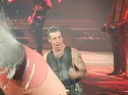 Mitte: Sänger Till Lindemann mit offenen Mund schaut nach oben und hält das Mikrofon in der linken Hand. Links unten im Vordergrund eine aktive Schaumkanone in orangener und hellroter Farbgebung. Die Eichel ist farblich hervorgehoben. Im Hintergrund stehen zwei Bandmitglieder mit Gitarre und Keyboard.