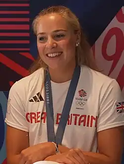 Rebecca Wilde lächelt im weißen T-Shirt der britischen Mannschaft. An einem blauen Band, das sie um den Hals trägt, ist ihre Olympiamedaille befestigt, die Wilde aber mit uhren Armen verdeckt.