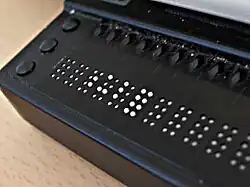 Die Nahaufnahme des linken Endes einer Braillezeile zeigt das schwarze Kunststoffgehäuse, links drei Funktionstasten, daneben einige 8-Punkt-Braille-Elemente. Diese zeigen mit ihren teilweise deutlich sichtbar angehobenen weißen Stößeln folgende Zeichen: zwei Leerzeichen, dann die Zeichen P56, P136, P123 und P12345678 und weitere Leerzeichen.