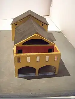 Das Modell zeigt ein detailgetreues architektonisches Miniaturgebäude, die Rekonstruktion von Strömbergs Theater. Es besteht aus zwei miteinander verbundenen Strukturen mit geneigten Dächern. Vorderer Teil des Gebäudes: Die Fassade ist gelb mit drei rechteckigen Fenstern. Im unteren Bereich befinden sich zwei große, bogenförmige Öffnungen. Der Innenraum ist sichtbar und in Rot gestaltet. Hinterer Teil des Gebäudes: Ähnlich im Design, aber aufgrund des Blickwinkels weniger prominent sichtbar. Dach: Dunkel gefärbt, möglicherweise schwarz oder dunkelgrau. Das Modell ist auf einer flachen, grauen Basis platziert, die den Untergrund symbolisiert.