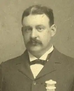 Mann mittleren Alters im dunklem Anzug, weißem Hemd und schwarzer Fliege mit angehefteten Brustorden von vorn, in die Kamera schauend ab Brustkorb stehend in schwarzweiß.