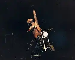 Halford mit freiem Oberkörper und zum Himmel gereckten Arm auf einem Motorrad