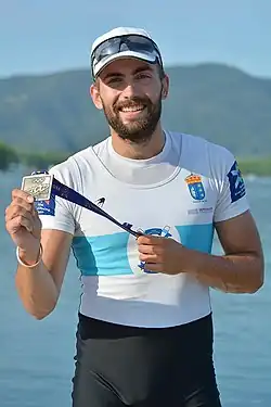 Rodrigo Conde trägt eine schwarze Hose, ein weißes Trikot mit blauem Querstreifen sowie eine weiße Kappe, seine Sonnenbrille ist auf die Kappe hochgeklappt. In den Händen hält er ein Band mit daran befestigter Medaille.