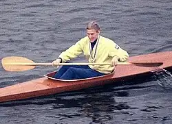 Rolf Peterson sitzt in einem schlanken braunen Rennkajak auf ruhigem Wasser. Er trägt einen gelben Pullover und eine Medaille um den Hals und hält ein Holzpaddel in beiden Händen.