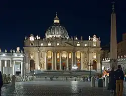 Frontale Nachtfotografie vom Petersdom mit wenigen Menschen auf dem Platz im Vordergrund. Der Dom mit seinem Säuleneingang und der Kuppel ist beleuchtet. An der rechten Seite ist ein dunkler Obelisk mit einer Kreuzspitze zu erkennen.