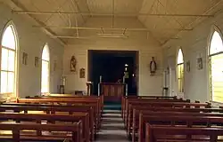 Das Foto eines Innenraums einer Holzkirche. Der Betrachter steht in der Kirche und blickt zwischen den beiden Bankreihen entlang auf den Holzaltar. Dieser steht in einer Nische. Rechts und links von der Nische befindet sich je eine Heiligenfigur an der Wand. Vor dem Altar steht ein Rednerpult. Die Wände und das Dach der Kirche sind weiß gestrichen und spärlich mit Bildern, deren Inhalt nicht erkennbar ist, dekoriert. Durch die schlichten Glasfenster auf der linken Seite scheint die Sonne in den Innenraum.