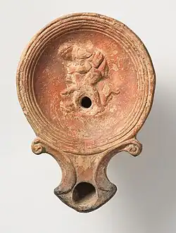 Im Spiegel der Lampe aus rotem Ton ist ein Mann dargestellt, der sich nach links über eine Ziege beugt, die er zwischen seinen Beinen fixiert und mit einem Messer in den Hals sticht, um sie zu schlachten. Zum Auffangen des Blutes steht ein Becken bereit.