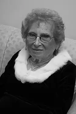 Das Schwarz-Weiß-Bild zeigt Tarlovsky de Roisinblit vor hellem Hintergrund sitzend auf einem hellen Sofa oder einer ähnlichen Sitzgelegenheit. Sie trägt ein dunkles Oberteil mit hellem Pelzsaum, Schmuck und eine Brille. Lächlend blickt sie in die Kamera.