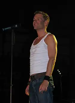 Peter Plate von Rosenstolz stehend auf der Bühne. Er trägt ein weißes Tanktop und eine blaue Jeans. Ein Gürtel ist um seine Taille gelegt, und er trägt ein schwarzes Armband am rechten sowie ein weiteres Armband am linken Handgelenk. Direkt neben Peter Plate steht ein Mikrofonständer mit einem befestigten Mikrofon. Der Hintergrund des Bildes ist dunkel gehalten und entstand während des Konzert in Leipzig 2006.