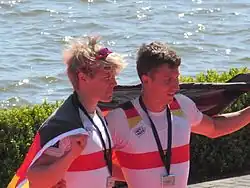 Die beiden Mainzer Ruderer im Nationaltrikot mit einer Silbermedaille auf der Brust halten hinter sich eine Deutschland-Fahne. Hinter ihnen ist eine Hecke und die Regattastrecke in Brandenburg erkennbar.