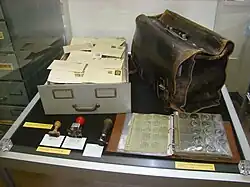 Ansicht einer Vitrine mit einer Schublade voller Briefe links, drei Stempeln davor und einem großen Lederkoffer rechts