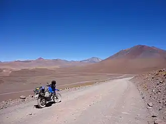 Der optisch unspektakuläre El Laco – Komplex gesehen aus etwa 15 km Entfernung. Nur der konische Gipfel fällt am Horizont über dem Motorrad auf.