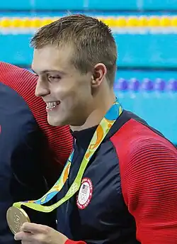 Ryan Held in blaurotem Trainingsanzug bei der Siegerehrung 2016, mit der rechten Hand zeigt er den Fotografen seine Goldmedaille.