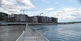 Foto einer Fußgängerbrücke, die über Wasser hinweg auf ein Gebiet führt, in dem mehrere Gebäude und Baukräne stehen