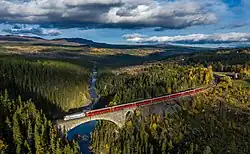 Foto eines Zugs auf einer steinernen Brücke, die in einer bewaldeten Landschaft einen Fluss überquert