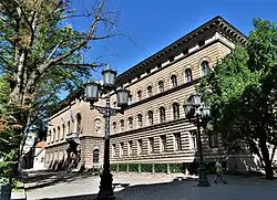 Eckansicht in Farbe von einem hellbraunen, dreigeschossigen Gebäude mit Rundbogenfenstern in den mittleren Geschossen. Im Vordergrund sind ein schwarz-weißer Transporter und zwei alte Straßenlaternen.