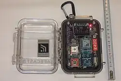 Das Safecast-Messgerät mit geöffnetem Deckel (zum Entnehmen der SD-Karte). Rechts Maßband zur Größeneinordnung