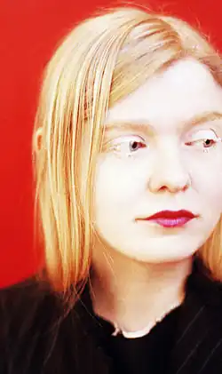 Sasa Hanten schaut nach rechts, hat langes, glattes, blondes Haar und rot geschminkte Lippen. Der Hintergrund ist rot.