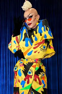 Velour steht auf einer Bühne, hinter ihr sind dunkelblaue Vorhänge zu sehen. Sie trägt roten Lippenstift, aufgemalte Augenbrauen, dunklen Lidschatten, falsche Wimpern, Wimperntusche, am linken Ohr einen gelben Ohrstecker, an dem eine Kette mit einer gelben Krone hängt, schwarze Handschuhe sowie eine Art gelben Morgenmantel, auf dem sich verschiedenfarbige Farbkleckse und schwarze Augen mit dunkelblauer Iris befinden. Mit der rechten Hand schiebt sie ihre Sonnenbrille mit roten Gläsern nach unten, während auf ihrem kahl rasierten Kopf eine weiße Plüschmaus sitzt.