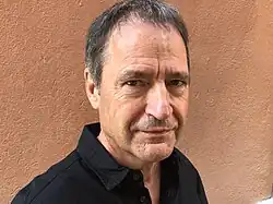 Walter Sigi Arnold (2021) trägt ein schwarzes Hemd. Der Hintergrund ist einfarbig und hat einen warmen, erdigen Farbton, der an Terrakotta erinnert. Er hat graues Haar und steht vor dieser schlichten Wand. Es sind keine weiteren Objekte oder visuellen Details im Bild enthalten.