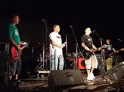 Vier jüngere und leger gekleidete Männer auf einer Bühne nebeneinander stehend mit Gitarren in der Hand und eine Person mit einem Mikrofon ins Publikum schauend.