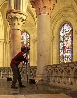 Seitliche Farbfotografie von einem Fotografen, der vorgelehnt durch eine Kamera mit Stativ blickt. Im Hintergrund sind Säulen und Kirchenfenster zu sehen.