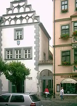 Altes Haus am Marktplatz, weiß verputzt, Stufengiebel an der Frontseite. Rechts daneben ist ein Durchgang zu einer anderen Straße zu sehen. Davor steht ein kleiner Baum.