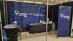 Zu sehen sind Banner mit der Aufschrift „smjg: Phantasie kennt keine Grenzen – Information auch nicht!“, ein Tisch mit Printmedien zum Mitnehmen und ein Tisch mit Kaffeemaschine, Bechern und Stofftier-Maskottchen