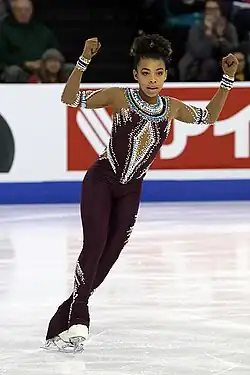 Starr Andrews, eine Schwarze junge Frau, trägt einen schwarzen, mit weißen Perlen besetzten Sportanzug (Unitard) zu ihren weißen Schlittschuhen. Sie gleitet auf der Außenkante des linken Schlittschuhs, den rechten Fuß vor den linken gestreckt. Die Arme hat sie erhoben, die Hände zu Fäusten geballt.