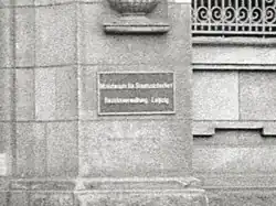 Schwarze geriffelte Metalltafel mit weißen Buchstaben, die in zwei Zeilen zeigen „Ministerium für Staatssicherheit“ und „Bezirksverwaltung Leipzig“