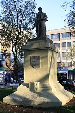 Die Statue Cannings in Santiago. Die Statue ist in einem kleinen umzäunten Bereich von Rasen umgeben. Sie hat eine Plakette mit einer Inschrift. Canning wird in einem Anzug dargestellt, seine rechte Hand geht zur Seite, seine linke Hand ruht auf einer halbhohen Säule. Das Gelände liegt einem bürgerlichen Viertel, im Hintergrund sind Geschäftsgebäude und Einkaufsläden zu erkennen.