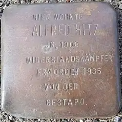 Abbildung des Stolperstein – Gedenksteins für Alfred Hitz, einer in Beton gegossenen Messingplatte von der größe eines Pflasterstein. Die Inschrift lautet: Hier wohnte Alfred Hitz JG.1908, Widerstandskämpfer, ermordet 1935 von der Gestapo.