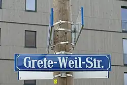 Blaues Straßenschild mit weißer Schrift "Grete-Weil-Str.". Das Schild ist an einem runden Holzpfahl angebracht, dahinter sieht man ein grauen Gebäude.