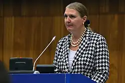 Das Bild zeigt le Jeune d’Allegeershecque in einer Naheinstellung vor dem braunen Hintergrund einer Holzvertäfelung. Sie steht an einem Rednerpult, trägt eine weiße Bluse und ein weiß-schwarz-kariertes Jackett, eine doppelreihige Perlenkette und Perlenohrringe. Ihre dunkelblonden Haare sind zu einem Zopf gebunden; sie blickt mit ernstem Gesichtsausdruck links am Betrachter vorbei.