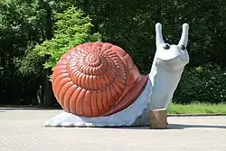 Eine sehr große Skulptur einer Schnecke, vor der ein Stück eines Baumstamms liegt, der klein erscheint; im Hintergrund Bäume