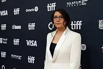 Sydney Freeland steht vor einer schwarzen Wand mit mehreren weißen Aufschriften. Sie trägt einen weißen, bis zur Brust aufgeknöpften Blazer, ein schwarzes Oberteil und eine dunkelbraune, runde Brille. Ihre schwarzen Haare trägt sie oben flach, unten strähnig bis zur Brust reichend.