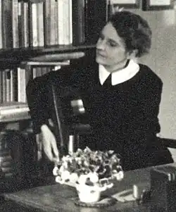 Sylvia Beach sitzt auf einem Stuhl, nach rechts gewendet, der rechte Arm über die Rückenlehne gelegt, als sei sie mit jemandem im Gespräch, der nicht im Bild zu sehen ist. Sie trägt das dunkle Haar hochgesteckt und ist schwarz gekleidet. Ein weißer Kragen bildet einen Kontrast dazu. Hinter ihr ist ein Regal mit Büchern, vor ihr auf dem Tisch ein Blumenstrauß zu sehen.