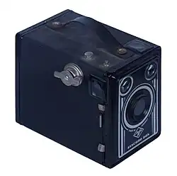 Farbfotografie einer Kastenkamera mit Tragegriff. An der Oberseite und der linken Seite ein Sucherfenster. Weiße Linien und das Logo der Agfa Synchro Box sind um das Objektiv gemalt.